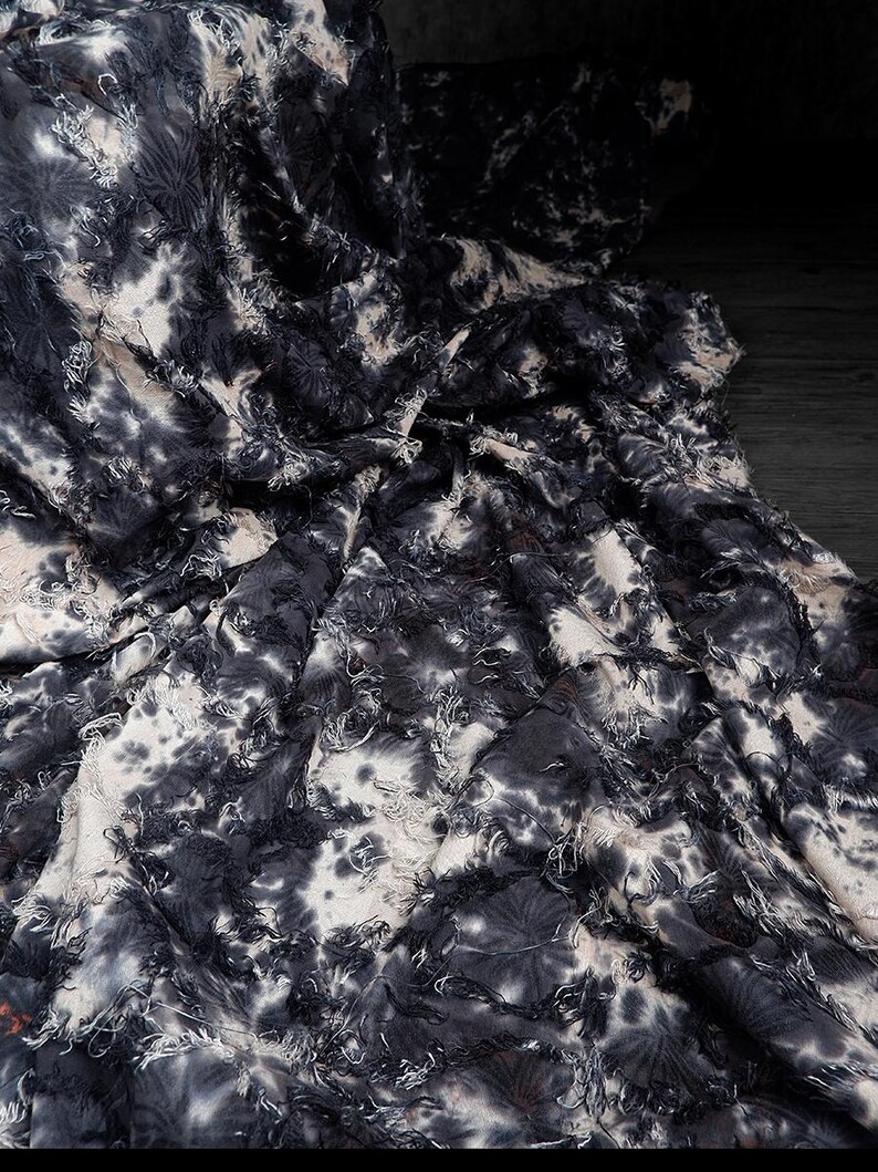 May include: Close-up of a fabric with a tie-dye pattern in shades of black, white, and gray. The fabric has a textured, frayed appearance, with visible threads and a crinkled texture. The overall effect is a dark, artistic textile.