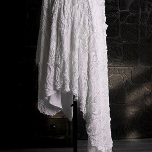 May include: White fabric with a textured, crinkled surface draped over a black stand. The fabric has a frayed edge and is displayed against a dark background.