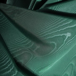 Può includere: Primo piano di un tessuto verde scuro con un motivo testurizzato e tessuto. Il tessuto è drappeggiato e piegato, creando linee morbide e fluide.