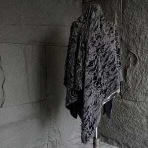 May include: A black, distressed, and tattered fabric garment draped over a wooden stand. The fabric has a unique, almost skeletal, pattern of holes and tears.