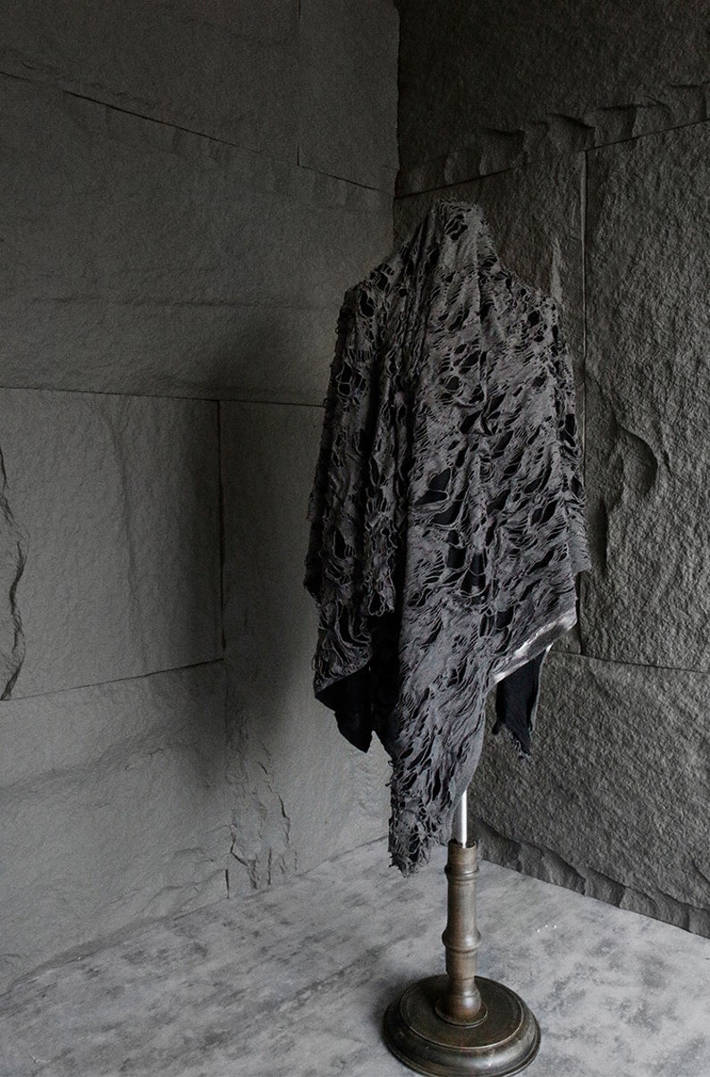 May include: A black, distressed, and tattered fabric garment draped over a wooden stand. The fabric has a unique, almost skeletal, pattern of holes and tears.