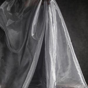 Może przedstawiać: Przezroczysta, srebrzysto-szara tkanina jest udrapowana, tworząc miękkie fałdy i refleksy. Tkanina wydaje się być trzymana, a światło odbija się od jej powierzchni. Tło jest ciemnoszare, co kontrastuje z połyskiem tkaniny.