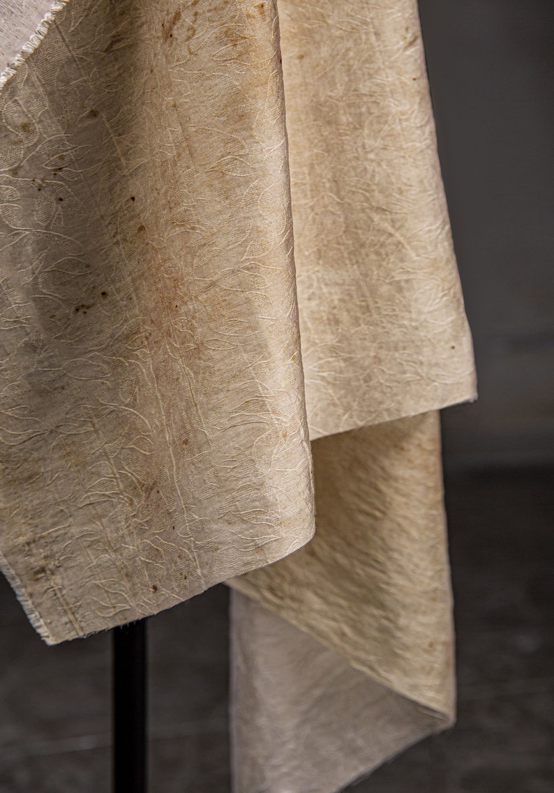 May include: A close-up of a cream-colored fabric with a textured pattern. The fabric is folded and draped over a black surface.
