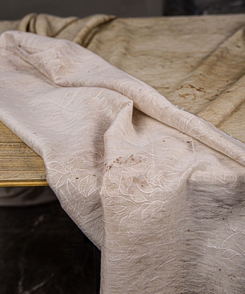 May include: A white tablecloth with a subtle floral pattern and brown stains. The tablecloth is folded and draped over a wooden surface.