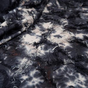 May include: Close-up of a fabric with a tie-dye pattern in shades of black, white, and gray. The fabric has a textured appearance with frayed edges, creating a layered effect. The textile is likely used for clothing or home decor.