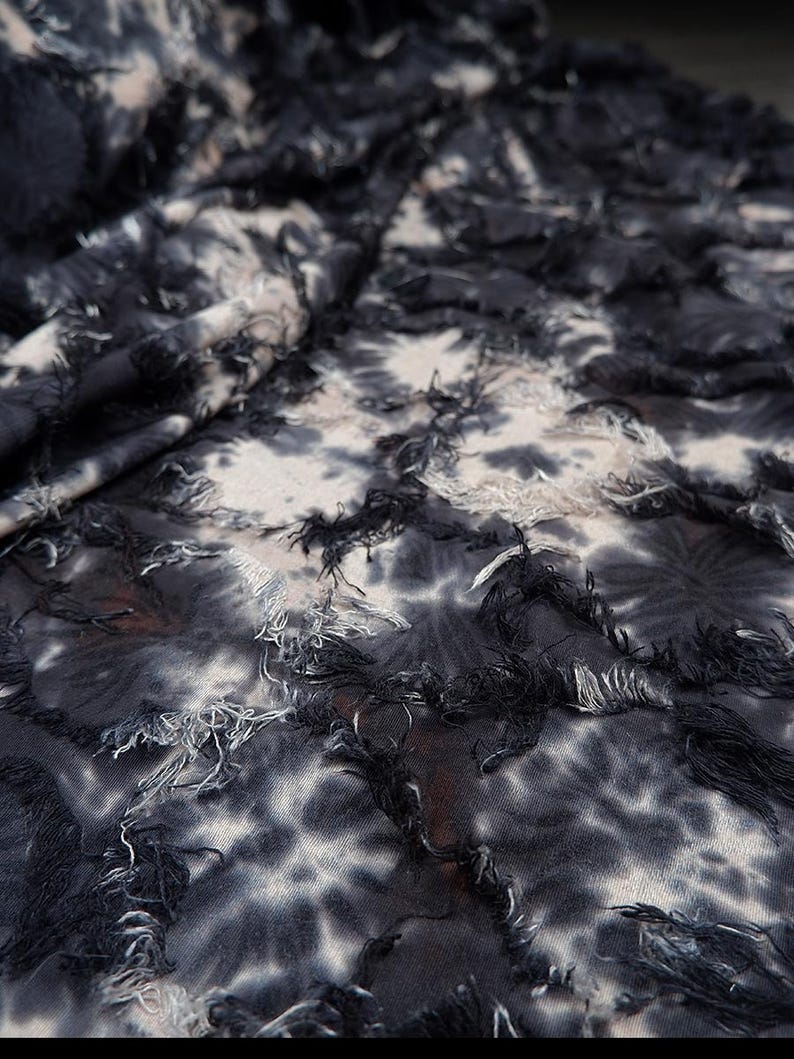 May include: Close-up of a fabric with a tie-dye pattern in shades of black, white, and gray. The fabric has a textured appearance with frayed edges, creating a layered effect. The textile is likely used for clothing or home decor.