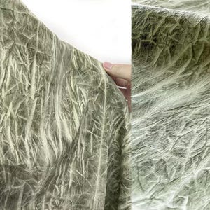 May include: Close-up of a textured fabric in shades of green and white. The fabric has a crinkled appearance, with a pattern of vertical and diagonal lines creating a unique, organic texture. The fabric is held at the edge.