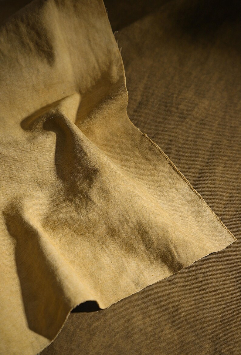 K&ouml;nnte beinhalten: Nahaufnahme eines St&uuml;cks beigen Stoffes mit rauer Textur. Der Stoff ist gefaltet, wodurch Schatten entstehen und die Textur hervorgehoben wird. Die Kanten des Stoffes sind sichtbar und zeigen eine Naht. Die Gesamtpalette ist erdig, mit Variationen von Beige und Braun.