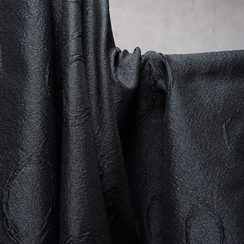 May include: Black fabric with a textured, circular pattern. The fabric is draped and folded, showing the texture and sheen.