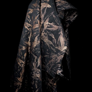 Puede incluir: Tela negra con un estampado floral bronce metálico. La tela está drapeada, mostrando la textura y el diseño. El patrón presenta hojas estilizadas y elementos botánicos, creando un textil lujoso.