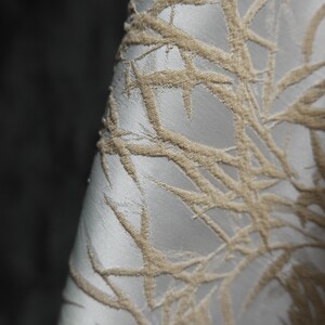 May include: Close-up of a white fabric with a raised, beige, geometric pattern. The pattern resembles branches or leaves.