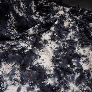 May include: Close-up of a fabric with a tie-dye pattern in shades of black, white, and gray. The fabric has a textured surface with frayed edges and a floral-like design. The fabric is draped, creating shadows and depth.