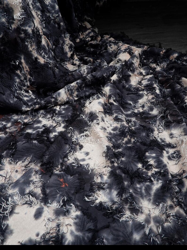 May include: Close-up of a fabric with a tie-dye pattern in shades of black, white, and gray. The fabric has a textured surface with frayed edges and a floral-like design. The fabric is draped, creating shadows and depth.