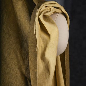 K&ouml;nnte beinhalten: Nahaufnahme eines drapierten, goldgelben Stoffes auf einer Mannequin-Schulter. Der Stoff hat ein strukturiertes Aussehen und einen Farbverlauf, der von einem dunkleren, olivfarbenen Farbton zu einem helleren, lebendigeren Gelb &uuml;bergeht. Die Kanten des Stoffes sind sichtbar.