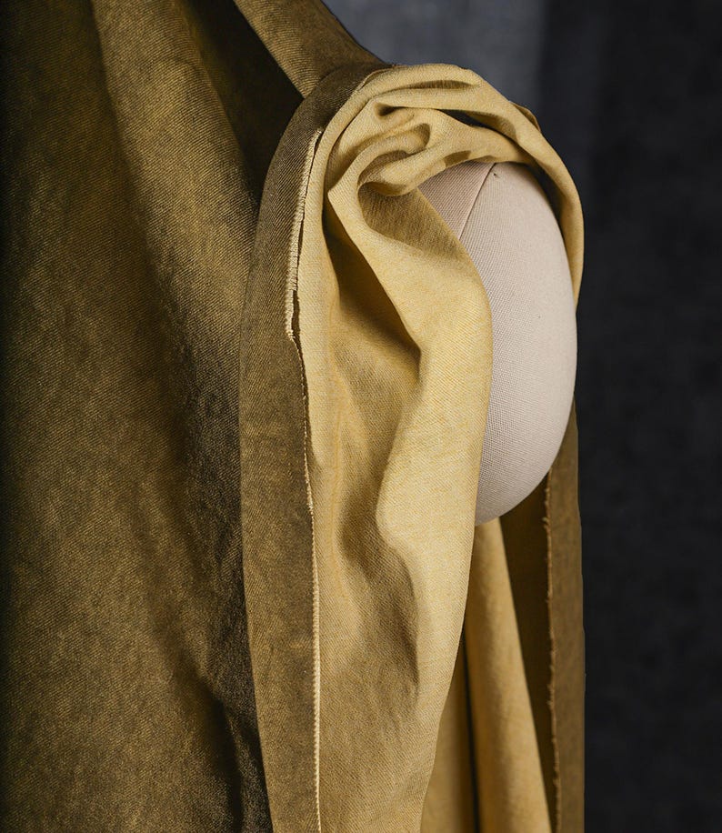 K&ouml;nnte beinhalten: Nahaufnahme eines drapierten, goldgelben Stoffes auf einer Mannequin-Schulter. Der Stoff hat ein strukturiertes Aussehen und einen Farbverlauf, der von einem dunkleren, olivfarbenen Farbton zu einem helleren, lebendigeren Gelb &uuml;bergeht. Die Kanten des Stoffes sind sichtbar.