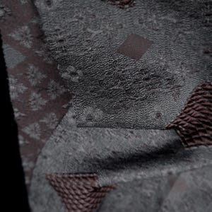 May include: Close-up of a textile with a patchwork design. The fabric features various textures and patterns, including floral motifs, diamond shapes, and woven sections in shades of gray and brown.
