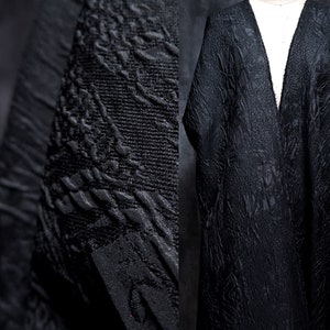 Puede incluir: Tela negra texturizada con un patrón sutil. La tela está drapeada sobre un maniquí, creando una prenda larga y fluida.