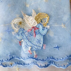 Puede incluir: Tres &aacute;ngeles bordados en vestidos azules con alas y halos dorados sobre una tela de tul azul claro con estrellas doradas.