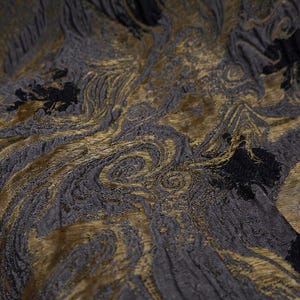 May include: Close-up of a textured fabric with a swirling pattern. The fabric features a combination of dark gray, black, and gold colors. The design has an abstract, organic appearance, with flowing lines and textured details.