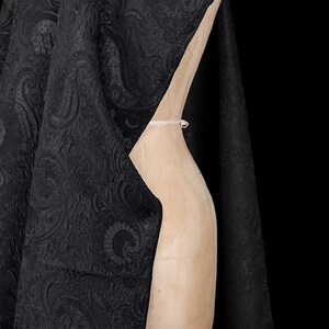 K&ouml;nnte beinhalten: Schwarzer Stoff mit Paisley-Muster, drapiert &uuml;ber einer h&ouml;lzernen Schneiderpuppe. Der Stoff hat eine strukturierte Oberfl&auml;che und ein sich wiederholendes, wirbelndes Design. Der Hintergrund ist einfarbig schwarz.