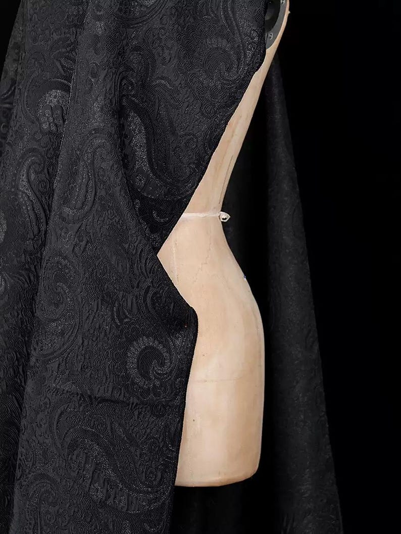 K&ouml;nnte beinhalten: Schwarzer Stoff mit Paisley-Muster, drapiert &uuml;ber einer h&ouml;lzernen Schneiderpuppe. Der Stoff hat eine strukturierte Oberfl&auml;che und ein sich wiederholendes, wirbelndes Design. Der Hintergrund ist einfarbig schwarz.