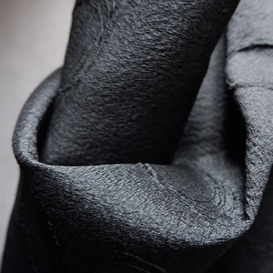 May include: Close-up of a black fabric with a textured surface. The fabric is folded and draped, showing the texture and drape of the material.