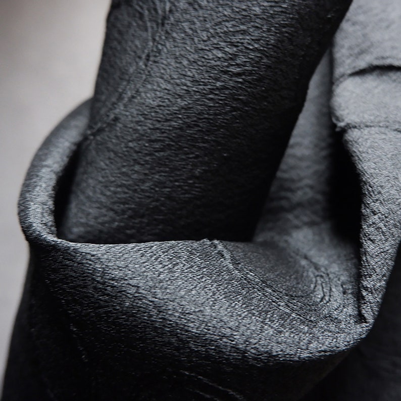 May include: Close-up of a black fabric with a textured surface. The fabric is folded and draped, showing the texture and drape of the material.