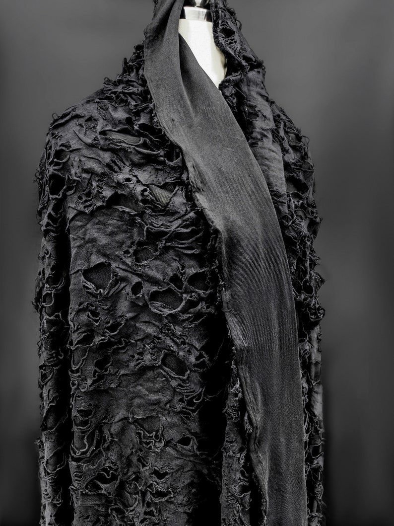 May include: A black, distressed fabric shawl with a frayed texture. The shawl is draped over a mannequin and has a long, flowing shape.