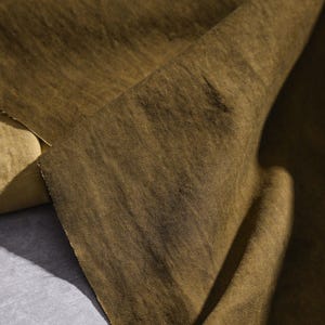 K&ouml;nnte beinhalten: Nahaufnahme eines gefalteten, olivgr&uuml;nen Stoffes. Der Stoff hat eine strukturierte Oberfl&auml;che mit sichtbaren diagonalen Linien. Die Farbe variiert leicht, wobei dunklere Bereiche einen Schatteneffekt erzeugen. Das Material scheint ein mittelschweres Textil zu sein.