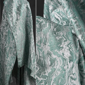 K&ouml;nnte beinhalten: Nahaufnahme eines teal- und wei&szlig;en Stoffes mit Marmormuster. Der Stoff ist &uuml;ber einem St&auml;nder drapiert und hat eine weiche, flie&szlig;ende Textur.