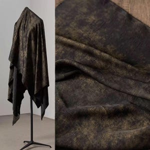 Op de afbeelding: Zwarte stof met een gouden metallic patroon gedrapeerd over een mannequin. De stof heeft een getextureerde uitstraling en wordt in twee aanzichten getoond, één gedrapeerd en de andere een close-up. De kleur van de stof is een donkere, gedempte tint.