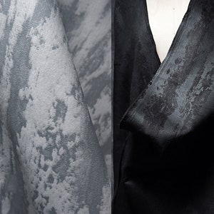 Puede incluir: Primer plano de una tela texturizada gris y blanca con un patrón sutil. La tela está drapeada sobre un maniquí, mostrando el drapeado y la textura del material.