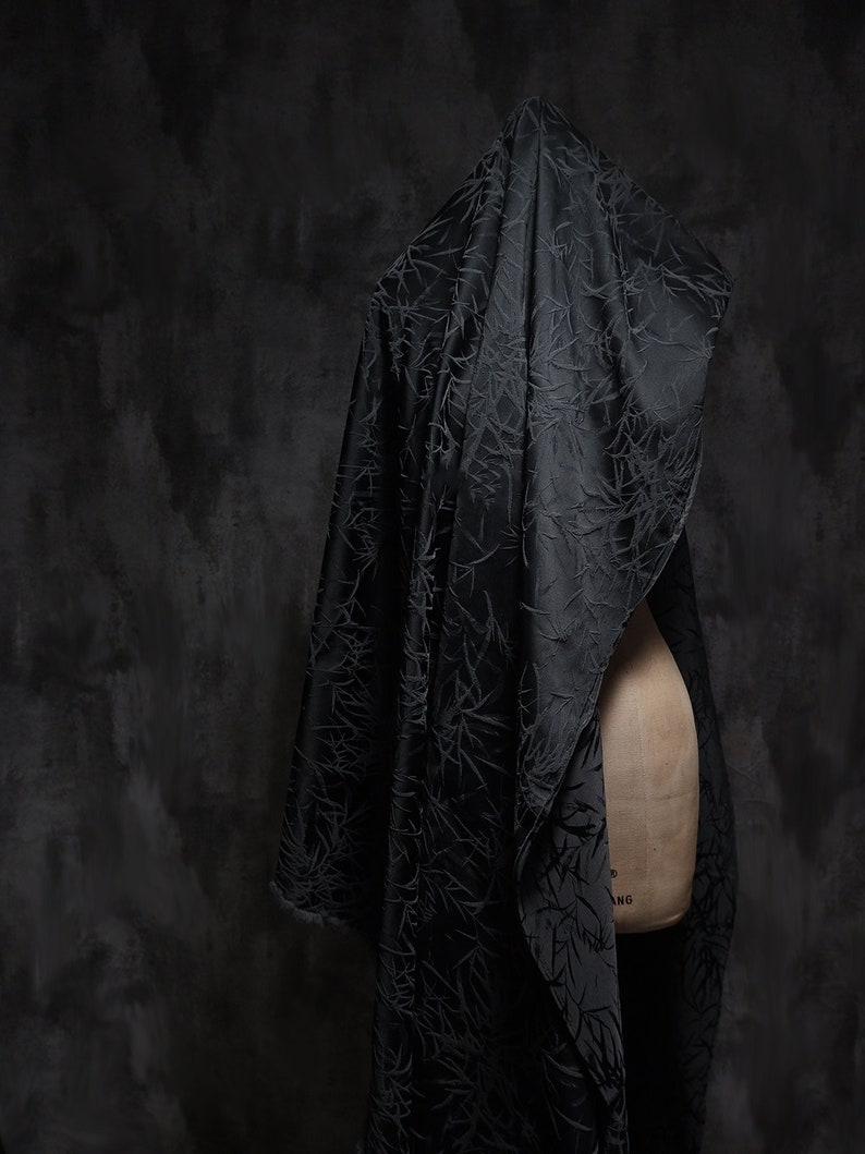 May include: A black fabric with a textured pattern draped over a dress form. The fabric has a subtle sheen and a delicate, almost floral, design.
