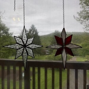 May include: Two star-shaped stained glass ornaments hanging from chains. One is clear, the other red and white. The background shows a green landscape. The ornaments are decorative and suitable for a window.