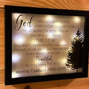 May include: A black framed light-up memorial plaque with a white background and a black silhouette of a pine tree. The text reads: "God saw you getting tired when a cure was not to be. He wrapped his arms around you and whispered "Come to me." You didn't deserve what you went through so He gave you needed rest. God's garden must be beautiful. He only takes the best. Martha Caldwell 1943-2015"