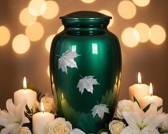 Puede incluir: Una urna verde con diseños de hojas de arce plateadas, colocada sobre una mesa con flores blancas y velas encendidas. El fondo presenta luces suaves y borrosas, creando una atmósfera serena y conmemorativa.