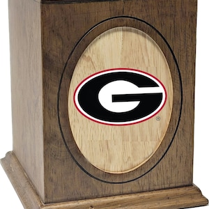 May include: A wooden urn with a dark brown finish. The front features an oval inset with the University of Georgia Bulldogs logo in black, red, and white. The urn has a rectangular shape with a decorative base and top.
