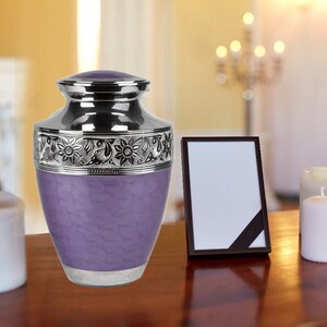 May include: A lavender cremation urn with a textured, scalloped pattern on the body. The urn features a polished silver lid and a silver band around the top decorated with an embossed floral motif, plus a silver base with an engraved border.