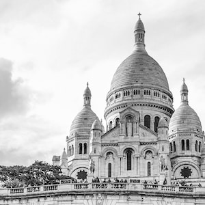 May include: A monochrome image of the Sacré-Cœur Basilica in Paris. The basilica has multiple domes and turrets, with detailed architecture. The sky is cloudy, and the building is constructed from white stone.