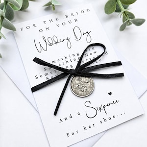 May include: A white card with black text reading For the bride on your wedding day, Something old, new, borrowed, blue, and a sixpence for her shoe. A black ribbon is tied around the card, holding a silver 1966 sixpence coin in the center.