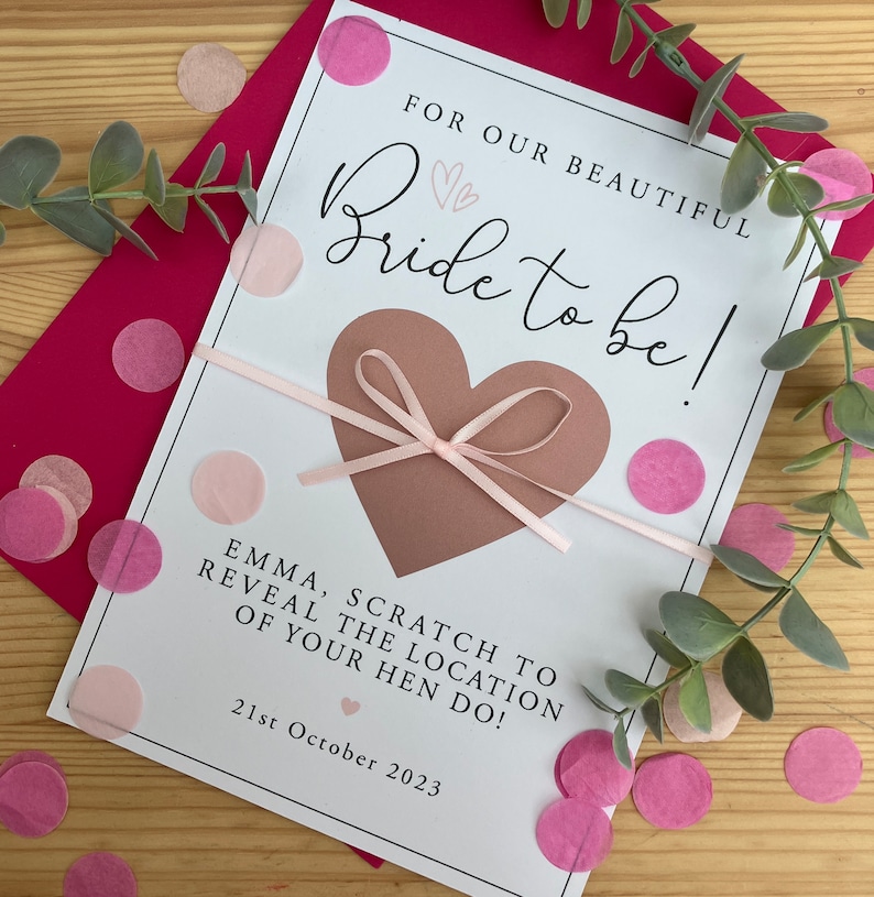 May include: A white card with a pink ribbon and a brown heart that says "FOR OUR BEAUTIFUL Bride to be!"  The card also says "EMMA, SCRATCH TO REVEAL THE LOCATION OF YOUR HEN DO!" and "21st October 2023".