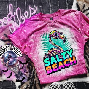May include: A pink t-shirt with a bleached design featuring a flamingo wearing sunglasses and the words "Salty Beach." The flamingo has a skeleton design. The shirt is on a zebra print surface.
