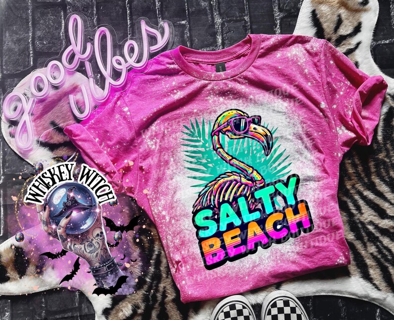 May include: A pink t-shirt with a bleached design featuring a flamingo wearing sunglasses and the words "Salty Beach." The flamingo has a skeleton design. The shirt is on a zebra print surface.