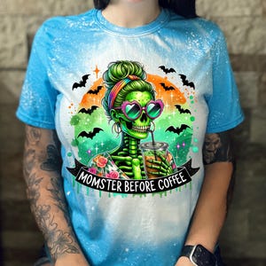 May include: A blue tie-dye t-shirt featuring a green skeleton with heart-shaped sunglasses, holding a coffee drink. The design includes bats, an orange and green background, and the text "MOMSTER BEFORE COFFEE" on a black banner.