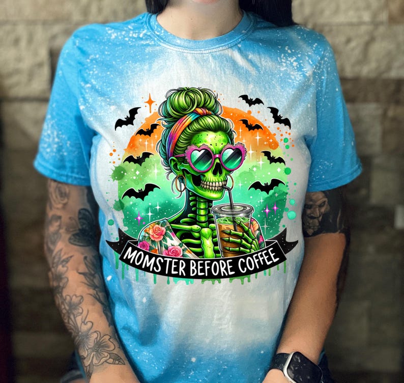 May include: A blue tie-dye t-shirt featuring a green skeleton with heart-shaped sunglasses, holding a coffee drink. The design includes bats, an orange and green background, and the text "MOMSTER BEFORE COFFEE" on a black banner.