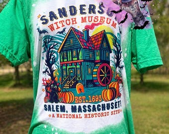 May include: Green t-shirt featuring a colourful graphic of the Sanders Witch Museum in Salem, Massachusetts. The design includes a house illustration, pumpkins, and the text "EST. 1693" and "A National Historic Site."
