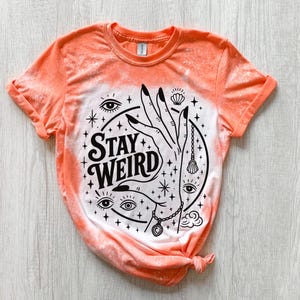 Handmade bleached styles shirt. Close-up of witchy Stay Weird shirt design with celestial artwork, mystical hands, stars, and bold black text