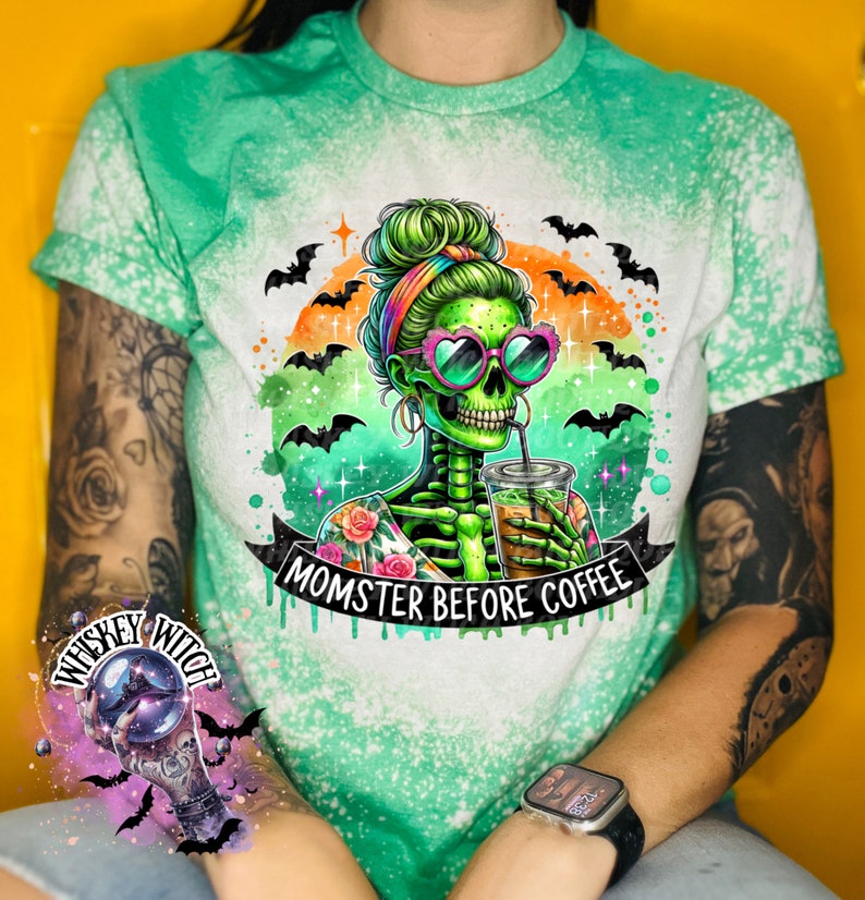 May include: Green and white tie-dye t-shirt featuring a skeleton graphic with heart-shaped sunglasses and the text "MOMSTER BEFORE COFFEE." The design includes bats, an orange and green background, and floral accents. The shirt has rolled-up sleeves.