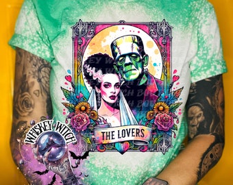 May include: A green t-shirt with a bleached effect features a colorful illustration of Frankenstein and his bride in a wedding scene. The text "The Lovers" is written in a banner above them. The image is framed with a decorative border.