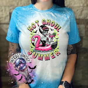 A sapphire blue  and bleach splattered effect  t-shirt with a graphic of a zombie woman in a flamingo pool float, holding a cocktail. The text "Hot Ghoul Summer" is printed on the shirt.